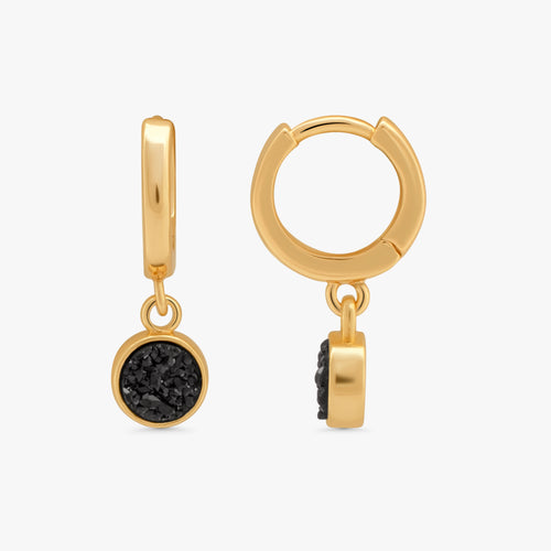A pair of gold huggie hoop earrings with a dangling round black textured stone charm, shown from both front and side angles on a light gray background.