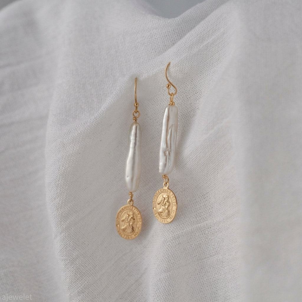 A pair of gold and white drop earrings displayed on a textured white fabric background. Each earring features a gold fishhook, a vertical white baroque pearl, and a dangling gold coin charm.