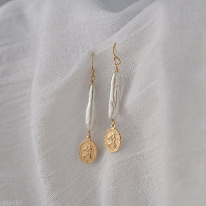 A pair of gold and white drop earrings displayed on a textured white fabric background. Each earring features a gold fishhook, a vertical white baroque pearl, and a dangling gold coin charm.