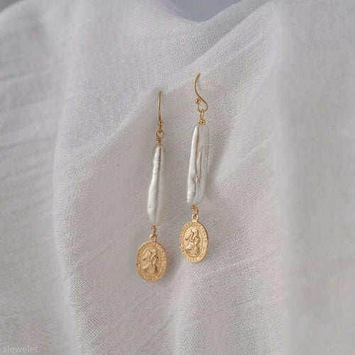 A pair of gold and white drop earrings displayed on a textured white fabric background. Each earring features a gold fishhook, a vertical white baroque pearl, and a dangling gold coin charm.