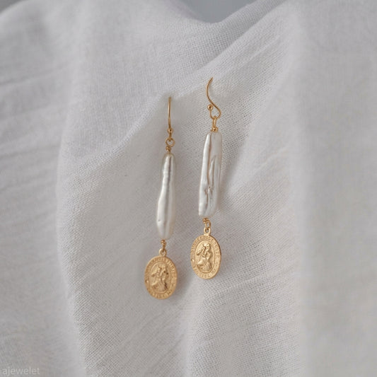 A pair of gold and white drop earrings displayed on a textured white fabric background. Each earring features a gold fishhook, a vertical white baroque pearl, and a dangling gold coin charm.