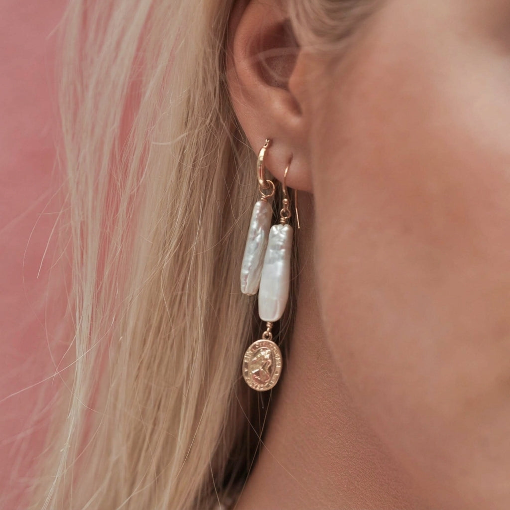 A close-up of a person's ear wearing a pair of dangling earrings. The earrings feature white baroque pearls and rose gold-toned metal hoops with a dangling round coin charm.