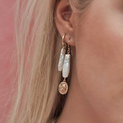 A close-up of a person's ear wearing a pair of dangling earrings. The earrings feature white baroque pearls and rose gold-toned metal hoops with a dangling round coin charm.