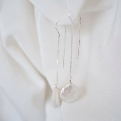 A pair of silver dangle earrings with irregular white baroque pearl pendants, shown hanging against a white satin fabric backdrop.