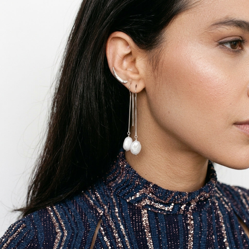 A close-up of a woman's ear wearing a pair of long silver drop earrings with three white baroque pearls, paired with a silver ear cuff.