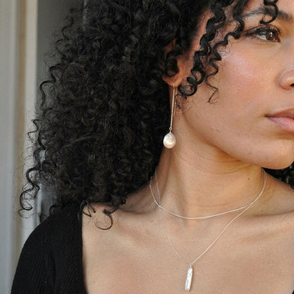 A close-up lifestyle shot of a woman with curly black hair wearing a silver-toned dangle earring with a white pearl, layered with a silver feather pendant necklace.