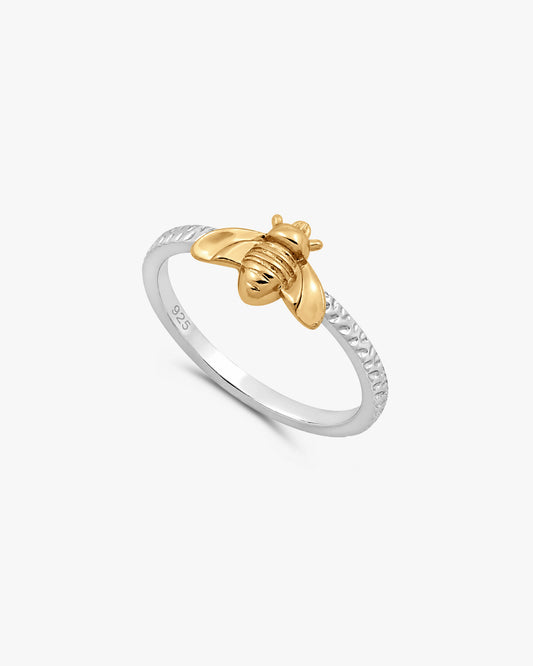 Two small bee-shaped rings on a white textured surface, one with a gold bee and a silver ring, with a blurred second pair in the background.