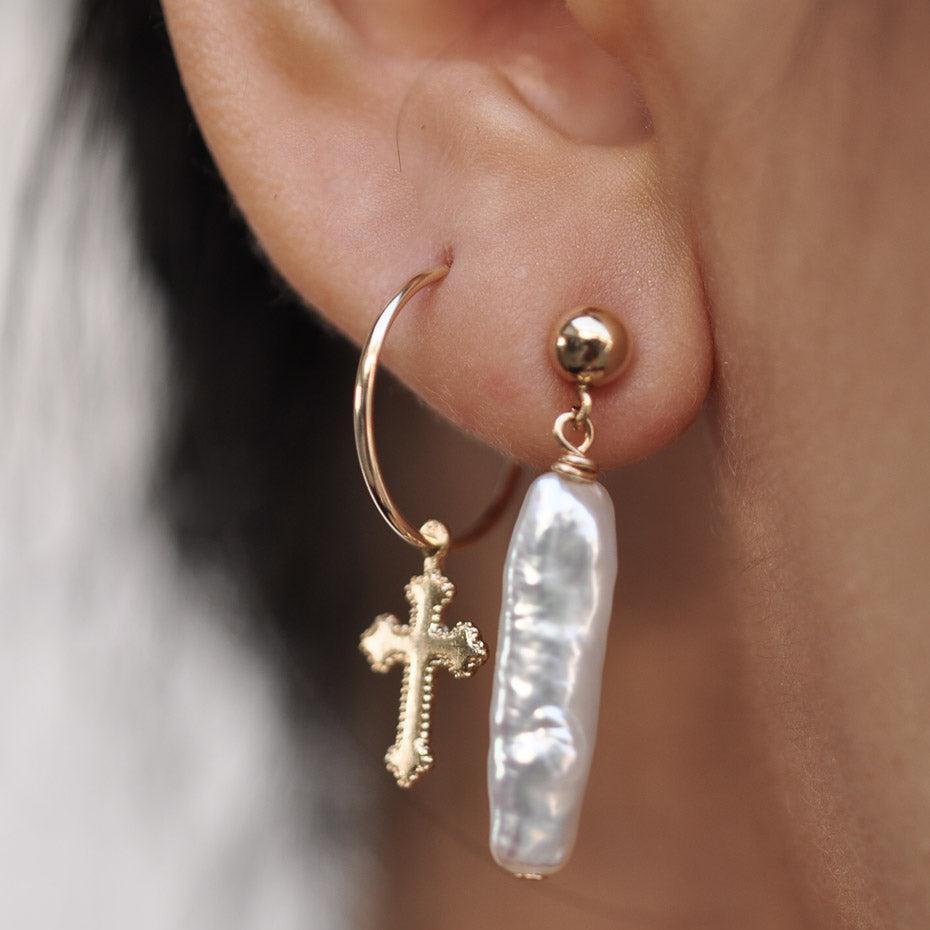 Woman wearing a 14k Solid Gold cross charm hoop earring alongside a long stick pearl drop stud.