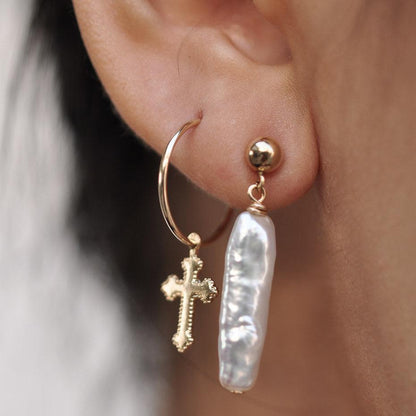 Woman wearing a 14k Solid Gold cross charm hoop earring alongside a long stick pearl drop stud.