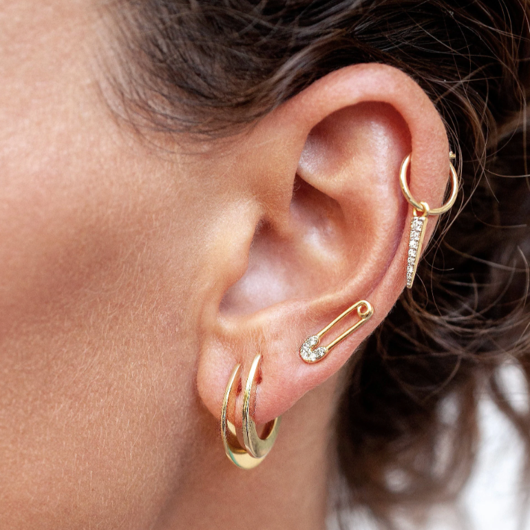 Woman wearing a gold crystal safety pin stud alongside a double hoop and spike dangle cartilage hoop.