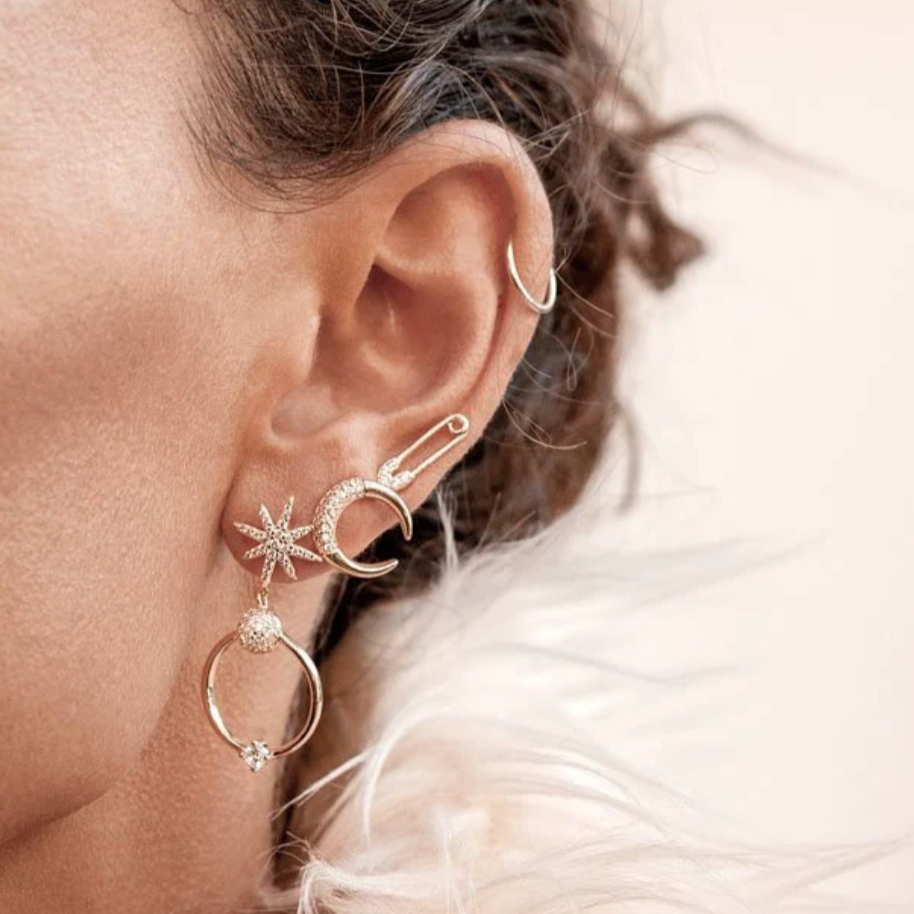 Woman wearing a gold safety pin stud, star drop earring, and crescent moon stud.