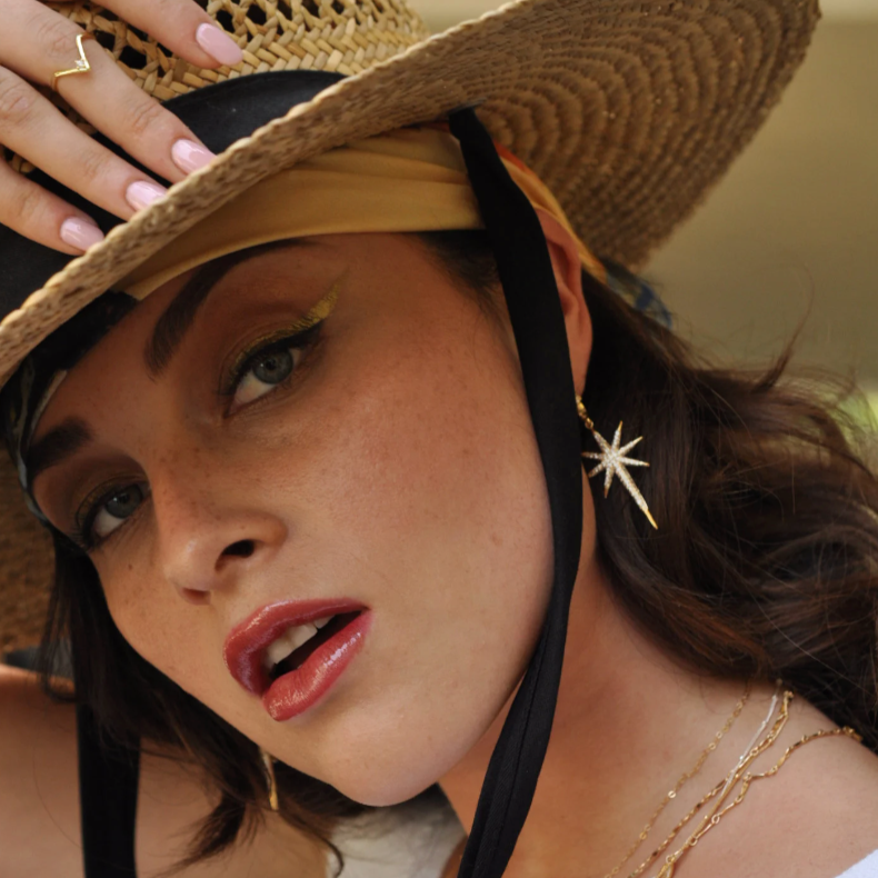 Woman wearing a sun hat and gold starburst drop earring with gold chain necklaces.