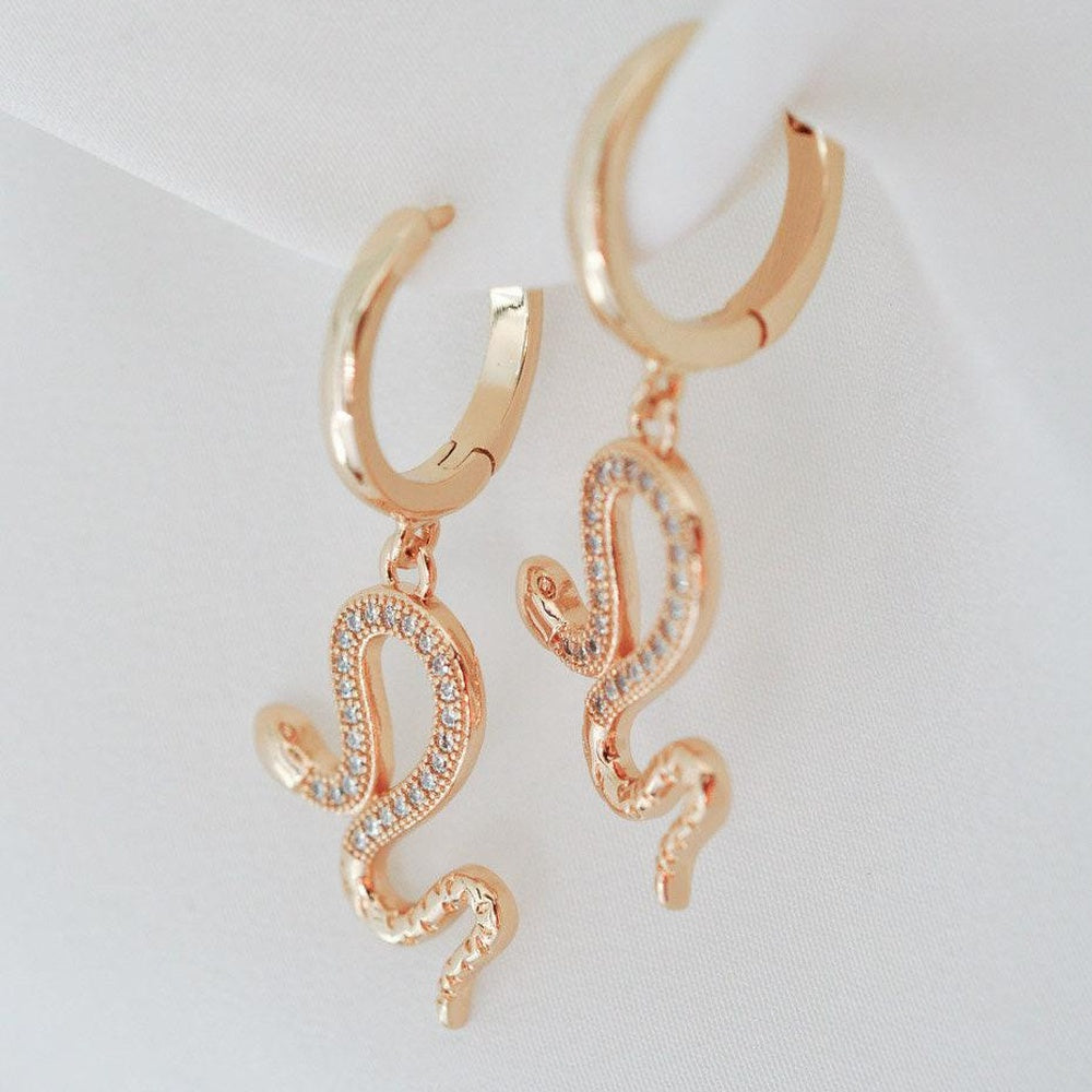 A pair of rose gold huggie hoop earrings with dangling snake charms embellished with small clear crystals, displayed on a white fabric background.