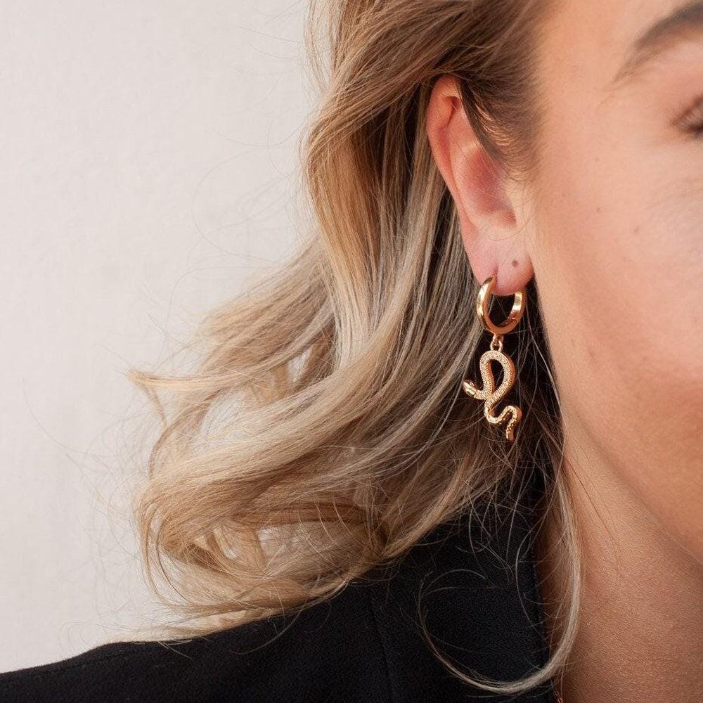 A close-up of a woman's ear wearing a gold hoop earring with a dangling stylized script letter 'S' charm, shown against a light neutral background.