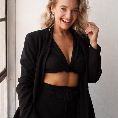 A woman smiling and wearing a black tailored blazer over a black cropped knit top and black high-waisted bottoms, standing indoors against a light wall near a window.