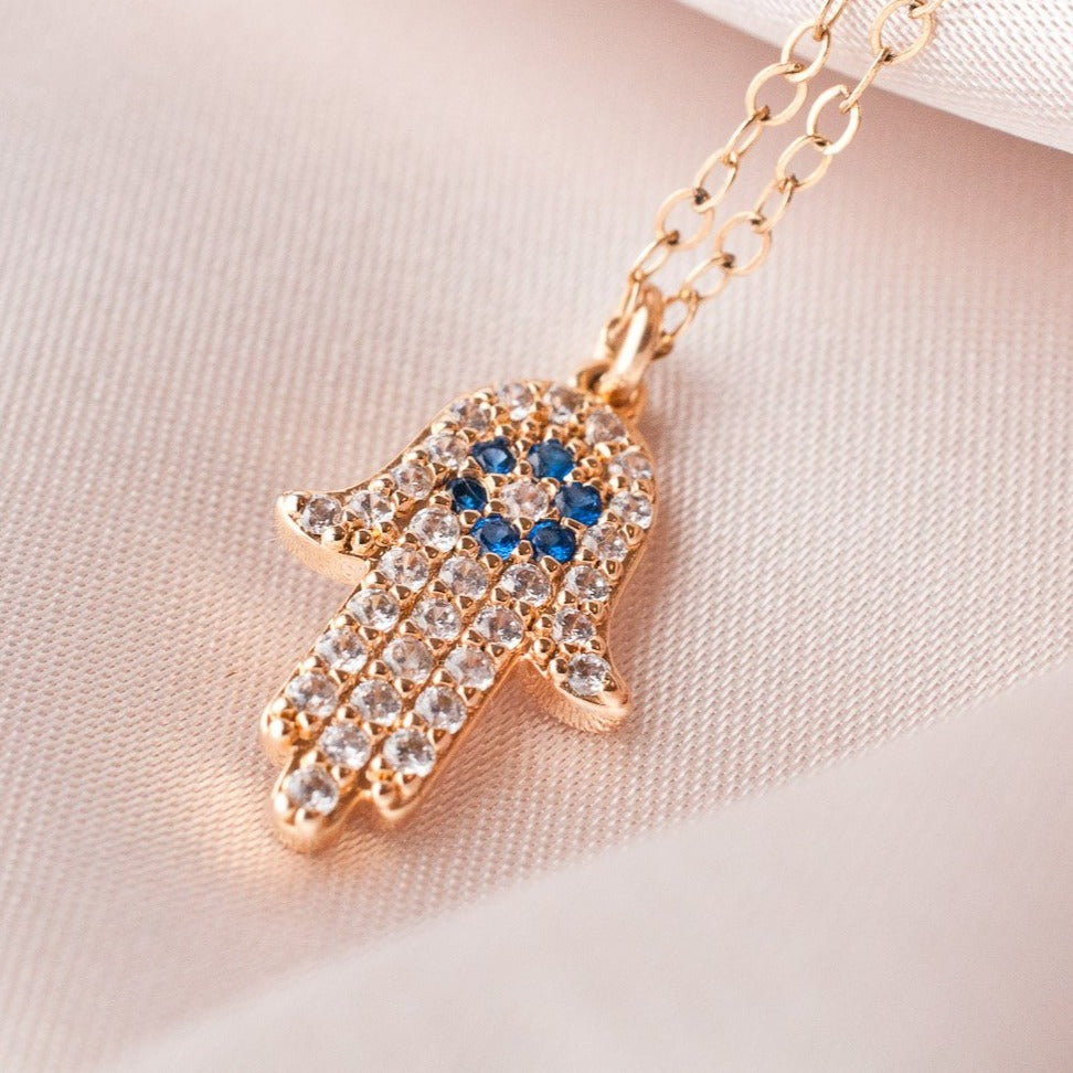 Asha Hamsa Hand 14k gold filled hamsa charm with sparkling clear cz crystals and blue stone center resting on soft white fabric
