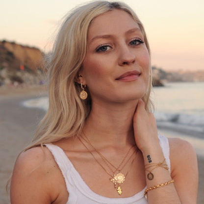 Blonde woman wearing Amora Mary 14k gold filled necklace layered with other gold jewelry at the beach