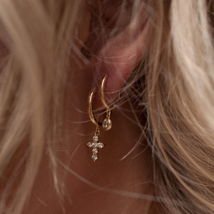Blonde woman wearing a 14k gold plated Lila solitaire crystal huggie drop earring stacked with a cross charm