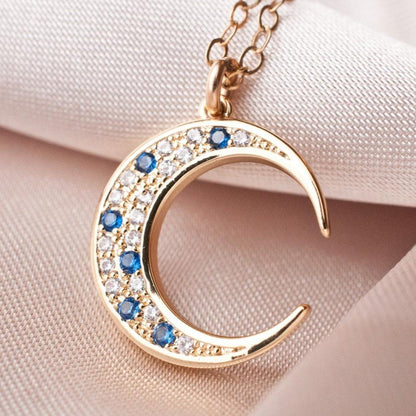Close-up of Elara Moon 14k gold filled crescent pendant with sapphire blue and clear crystals on silk fabric