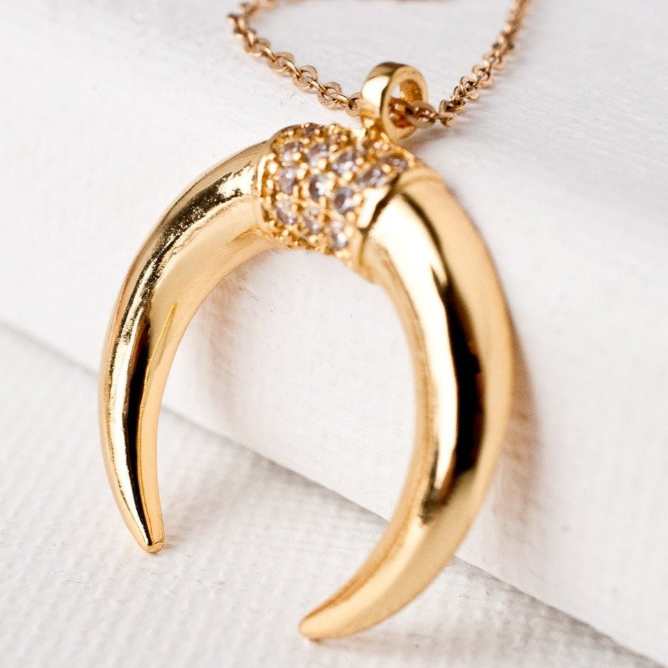 Close-up of Fara Double Horn Tusk 14k gold filled crescent pendant with crystal accents on white surface