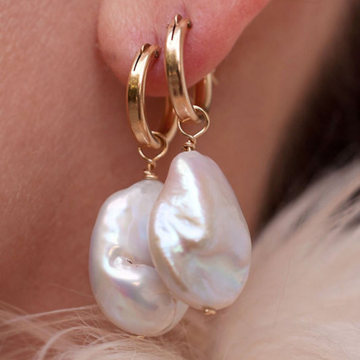 A close-up of a person wearing a pair of gold-colored hoop earrings with white baroque keshi pearl accents.
