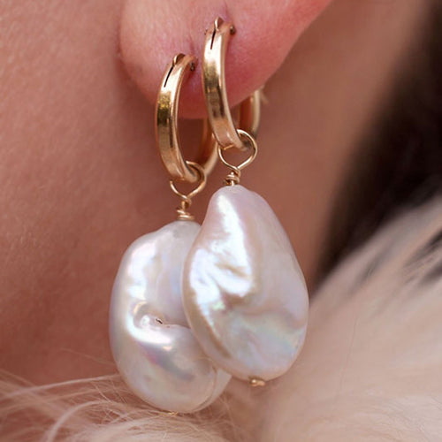 A close-up of a person wearing a pair of gold-colored hoop earrings with white baroque keshi pearl accents.