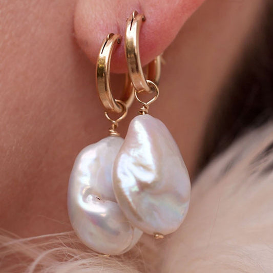 A close-up of a person wearing a pair of gold-colored hoop earrings with white baroque keshi pearl accents.