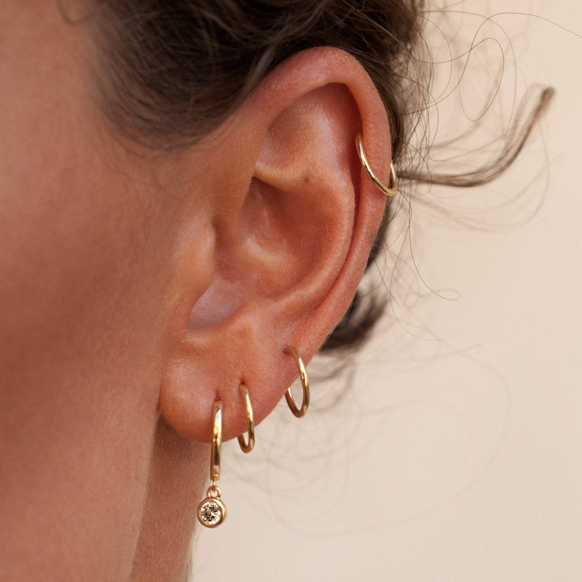Close up of a woman wearing 14k gold plated Lila solitaire crystal huggie drop earrings in a layered ear stack