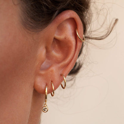 Close up of a woman wearing 14k gold plated Lila solitaire crystal huggie drop earrings in a layered ear stack