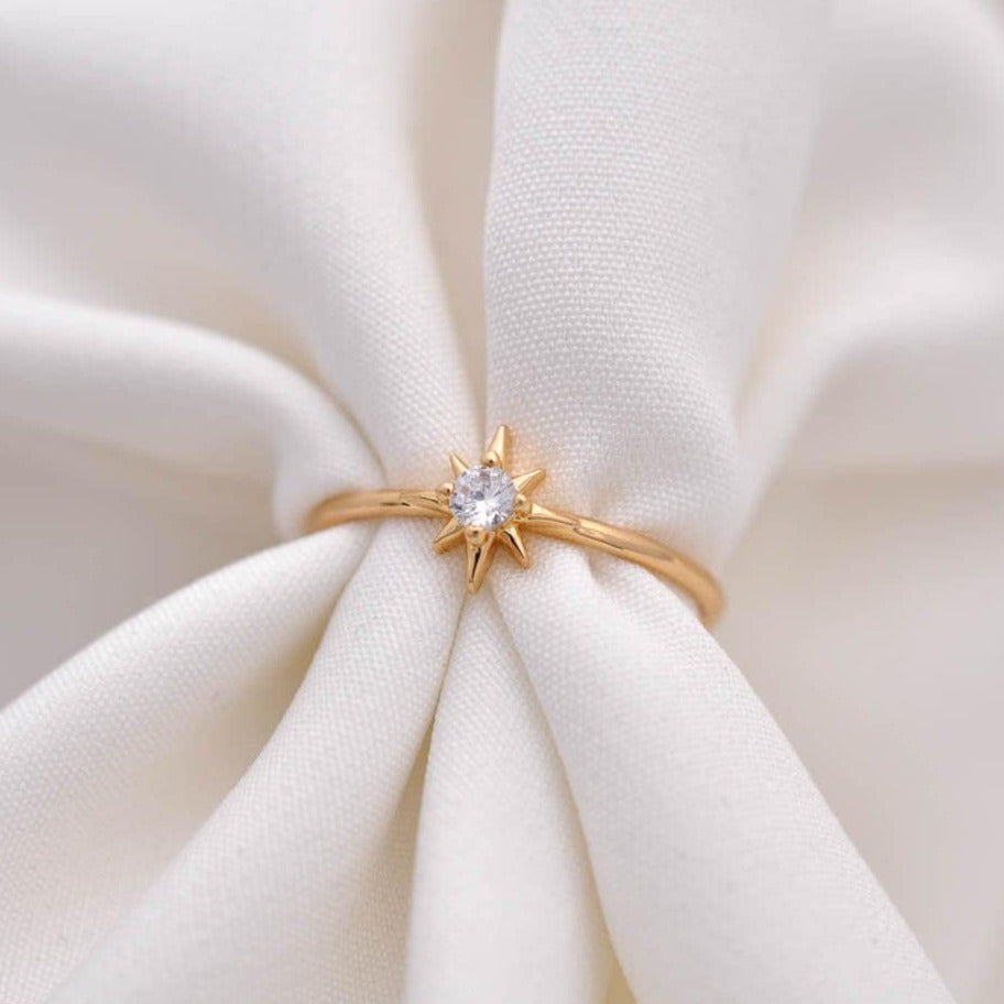 Gold starburst ring with a clear crystal solitaire on white draped fabric.