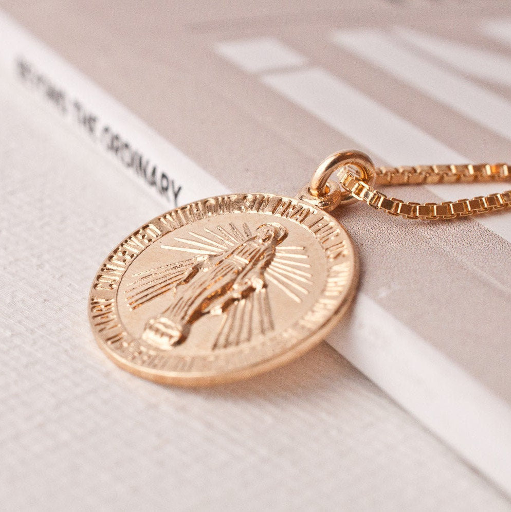 Macro detail of Annika Mary 14k gold plated Miraculous Mary medallion resting on white book surface