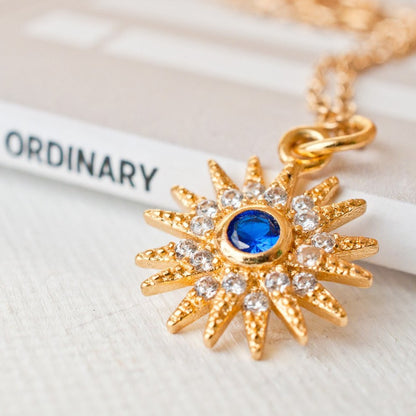 Macro detail shot of Sunniva Sun Ray & Starburst 14k gold filled pendant with blue and clear cz crystals on white surface