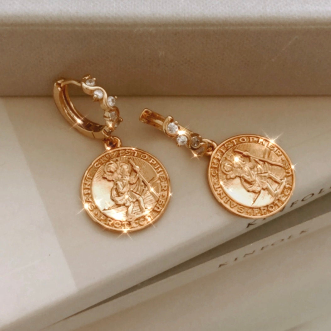 Pair of women's 14k Gold Plated Hallow Saint Christopher Hoops with coin charms resting on a white surface.