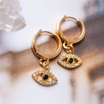 Pair of women's 14k Gold Plated Mira Evil Eye charm hoop earrings styled on a textured background near quartz crystals.