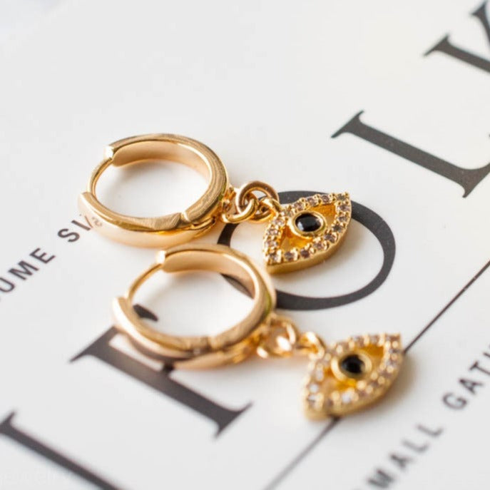 Pair of women's 14k Gold Plated Mira Evil Eye huggie hoop earrings resting flat on a magazine page.