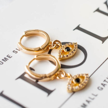 Pair of women's 14k Gold Plated Mira Evil Eye huggie hoop earrings resting flat on a magazine page.