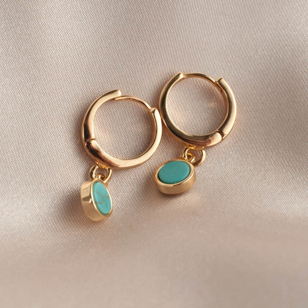 Pair of women's 14k Gold Plated Riva Turquoise Charm Hoop Earrings resting flat on soft beige satin cloth.