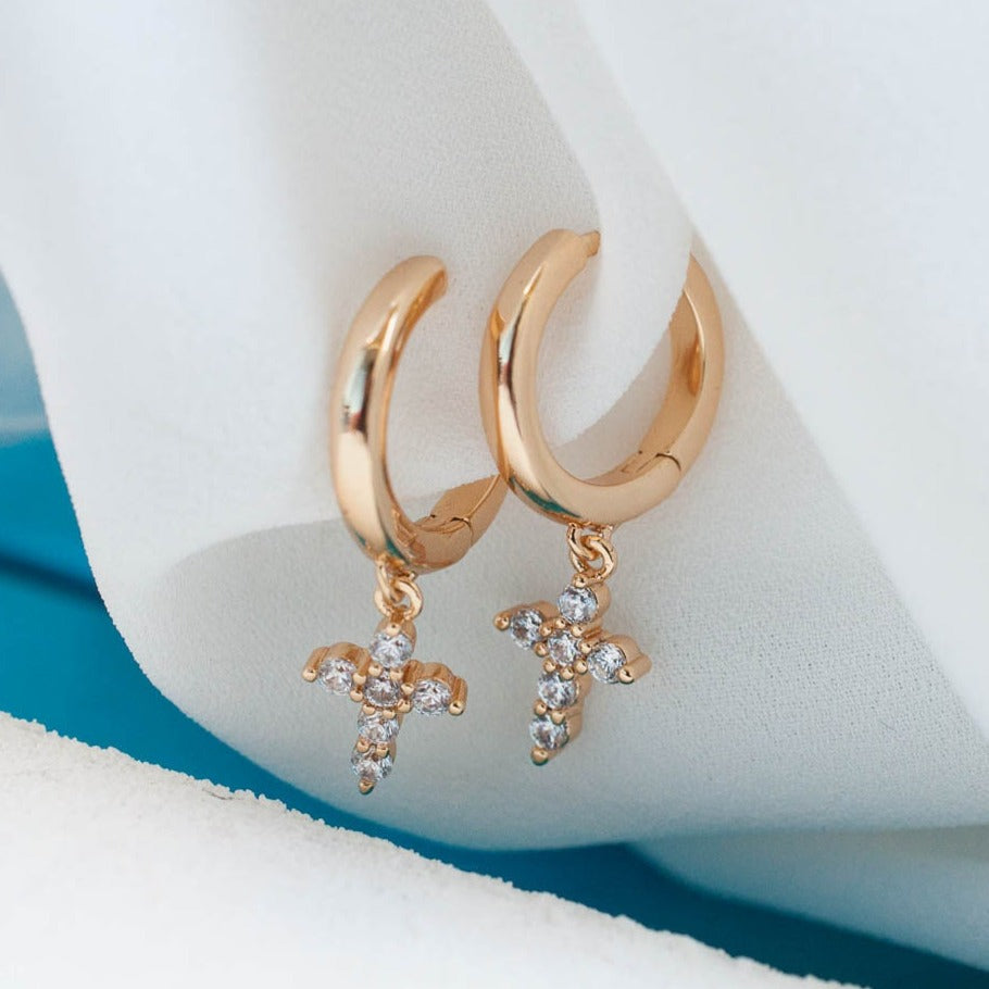 Pair of women's 14k Gold Plated Solid Mini Cross Hoops resting on white fabric against a turquoise background.