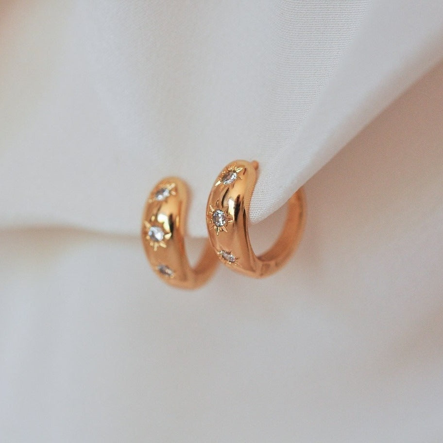 Pair of women's 14k Gold Plated chunky huggie hoop earrings with starburst crystal set details clipped on white fabric.