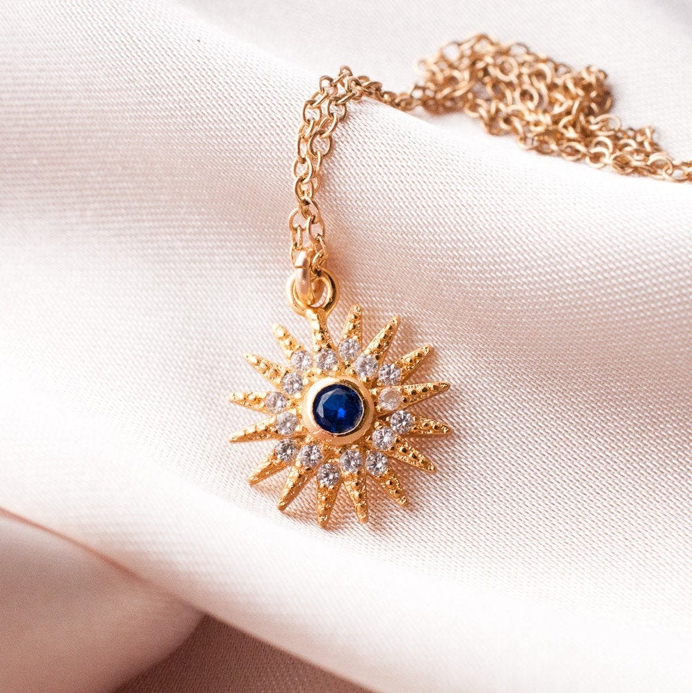 Sunniva Sun Ray & Starburst 14k gold filled necklace with blue crystal center displayed on soft white fabric