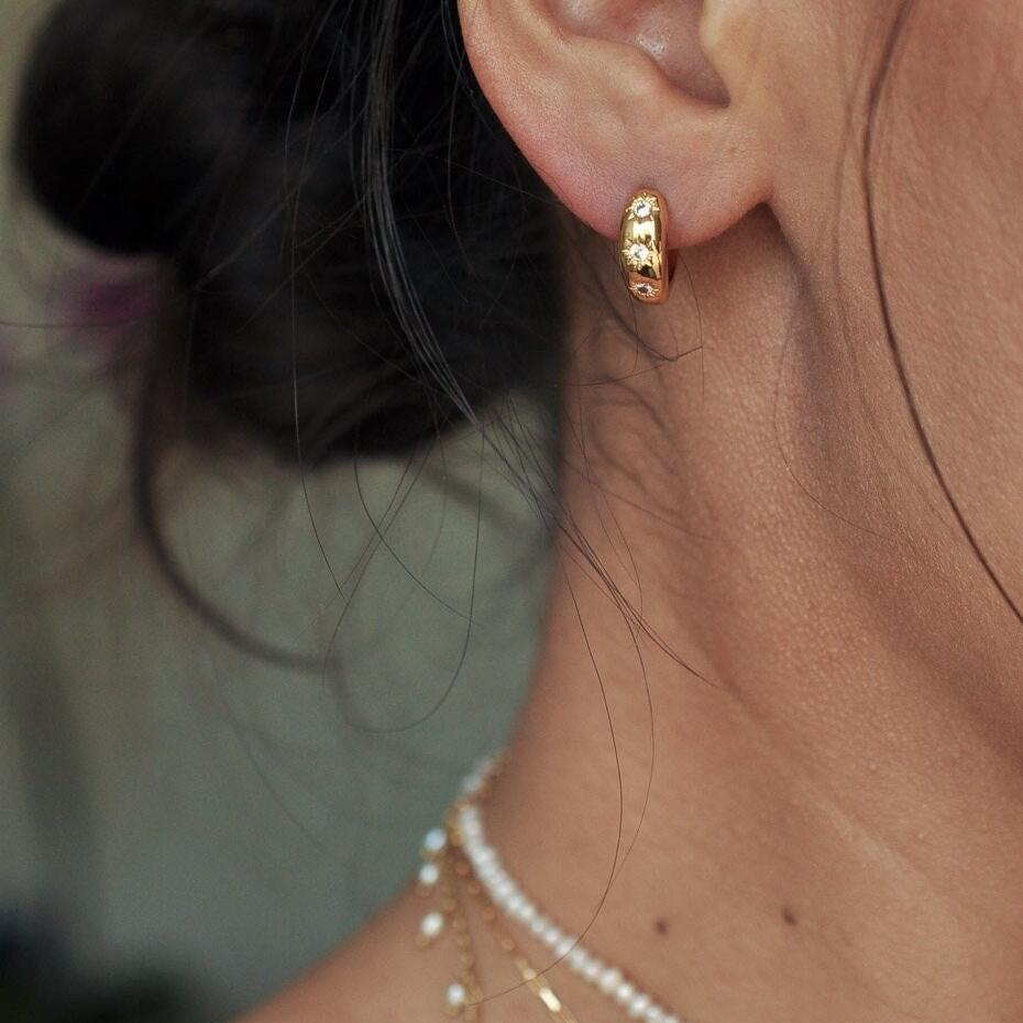 Woman with dark hair tied back wearing a small 14k Gold Plated starburst huggie hoop earring and pearl necklace.
