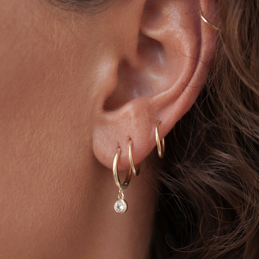 Woman wearing gold huggie solitaire diamond zirconia earrings with sleeper hoop earrings, she has a curly hair and tan skin.